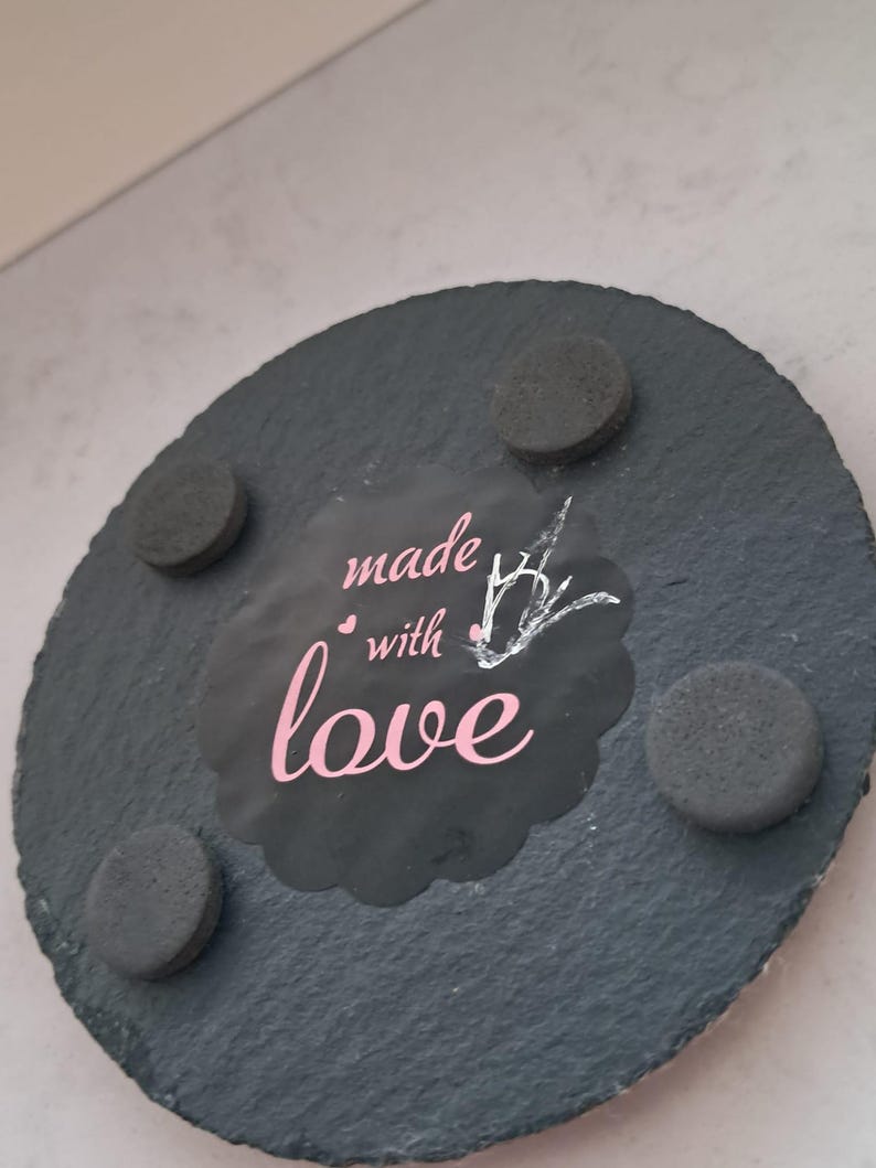 May include: A round, dark gray slate coaster with a textured surface. The center features a black scalloped label with the pink text "made with love" and a small white line-art drawing of a bird. Four circular black foam feet are attached.