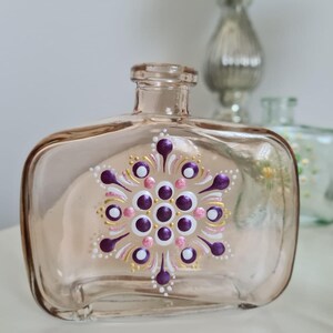 Op de afbeelding: Een heldere glazen fles met een roze tint en een geschilderd mandala-ontwerp. De mandala heeft paarse, witte, roze en gouden accenten.