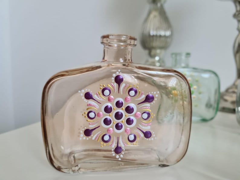 Op de afbeelding: Een heldere glazen fles met een roze tint en een geschilderd mandala-ontwerp. De mandala heeft paarse, witte, roze en gouden accenten.