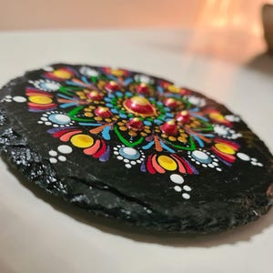 May include: A round black slate coaster featuring a hand-painted mandala design. The artwork consists of intricate, colorful dot patterns in shades of yellow, red, blue, green, and white, arranged in a symmetrical radial formation on the dark stone.