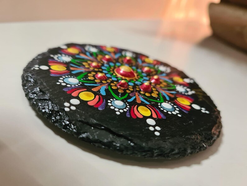 May include: A round black slate coaster featuring a hand-painted mandala design. The artwork consists of intricate, colorful dot patterns in shades of yellow, red, blue, green, and white, arranged in a symmetrical radial formation on the dark stone.