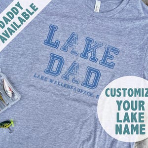 Könnte beinhalten: Ein graues T-Shirt mit blauem Text, der "LAKE DAD" und "LAKE WALLENFAUPACK, PA" lautet. Ein weißer Kreis mit grünem Text sagt "CUSTOMIZE YOUR LAKE NAME".