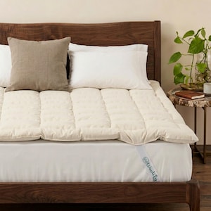 Op de afbeelding: Een bed met een crèmekleurige matrasbeschermer, witte kussens en een bruin kussen. Het bedframe is van donker hout. Een klein bijzettafeltje met een plant en een notitieboek is zichtbaar.