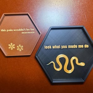 May include: Two hexagonal coasters. One is brown with the text "this pain wouldn't be for evermore" and snowflake designs. The other is black with the text "look what you made me do" and a golden snake design.