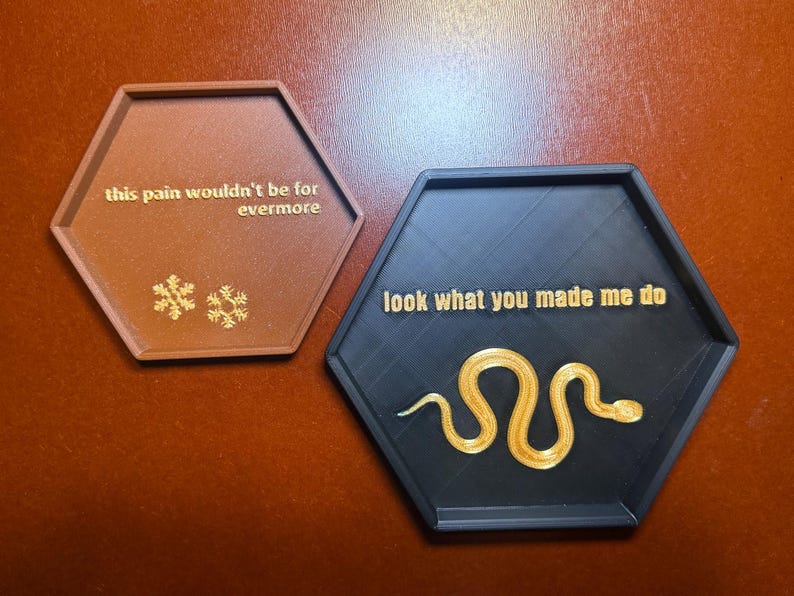 May include: Two hexagonal coasters. One is brown with the text "this pain wouldn't be for evermore" and snowflake designs. The other is black with the text "look what you made me do" and a golden snake design.