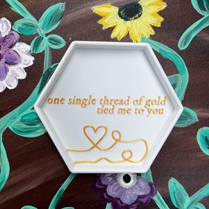 May include: White hexagonal ceramic trinket dish with a gold inscription that reads "one single thread of gold tied me to you". The inscription is accompanied by a gold heart design.