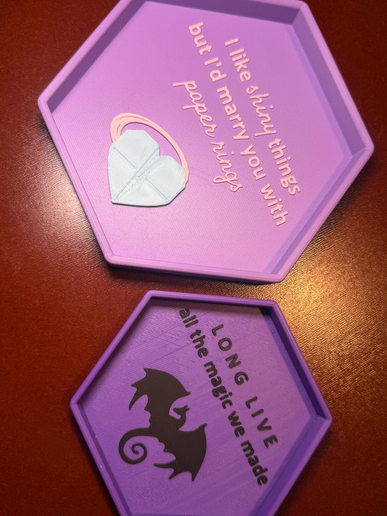 May include: Two hexagonal purple trays. The top tray has the text "I like shiny things but I'd marry you with paper rings" and a heart-shaped accessory. The bottom tray has a dragon silhouette and the text "LONG LIVE all the magic we made."