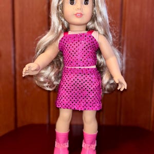 May include: A pink sequined dress and matching top outfit for a doll. The doll has long blonde hair and brown eyes. The doll is wearing pink boots with white soles.