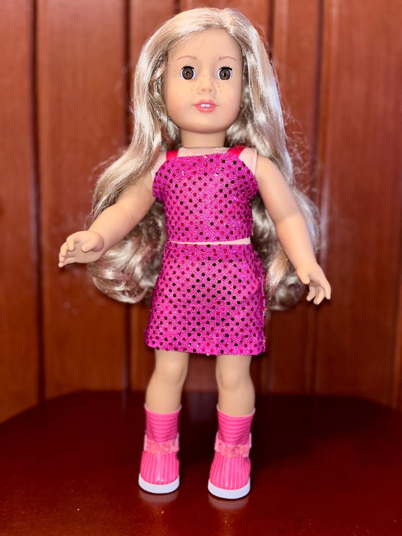 May include: A pink sequined dress and matching top outfit for a doll. The doll has long blonde hair and brown eyes. The doll is wearing pink boots with white soles.