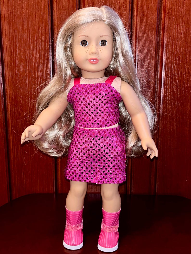 May include: A doll wearing a pink sequined dress and pink boots. The doll has long blonde hair and brown eyes.