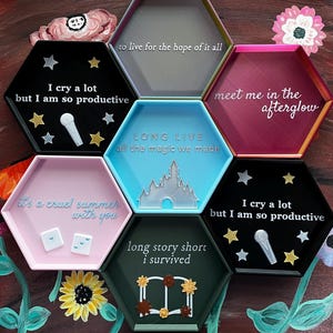 May include: A collection of hexagonal trays in various colors, each with a unique design and text. The trays feature phrases like "I cry a lot but I am so productive" and "Long Live all the magic we made". Some trays have star and microphone accents.