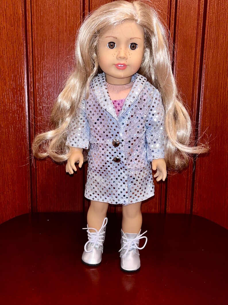 May include: A silver sequined jacket and dress outfit for a doll. The doll has blonde hair and brown eyes. The outfit has a button closure and a collar.