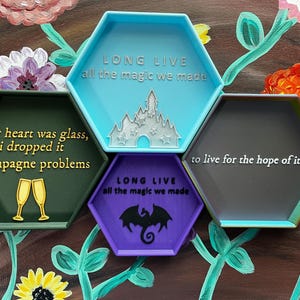 May include: A collection of colorful, hexagonal trays with inspirational quotes and imagery. The trays are in shades of green, blue, purple, and gray, with gold and white text. The image includes floral artwork in the background.