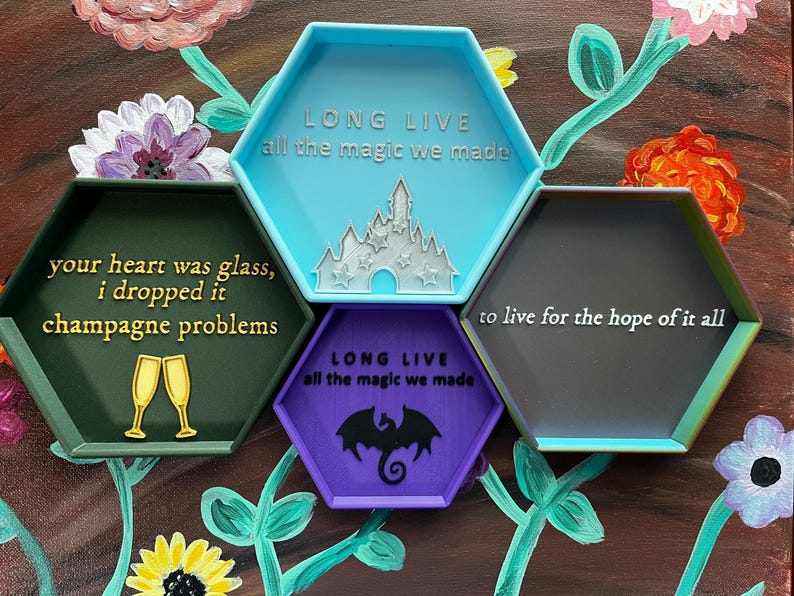 May include: A collection of colorful, hexagonal trays with inspirational quotes and imagery. The trays are in shades of green, blue, purple, and gray, with gold and white text. The image includes floral artwork in the background.