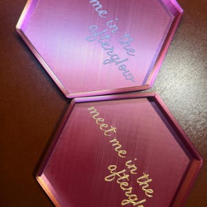 May include: Two hexagonal trays in shades of pink and magenta. The trays have raised edges and feature the text "meet me in the afterglow" in a cursive font. The trays are likely for decorative or organizational purposes.