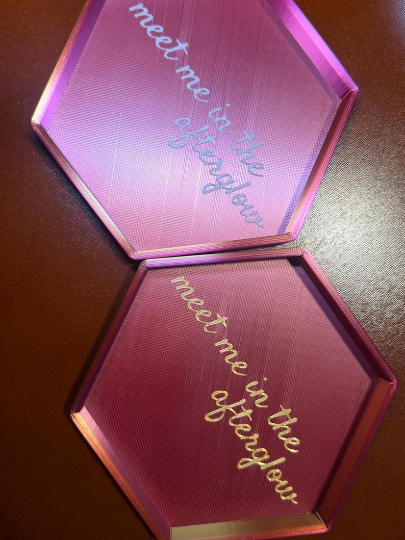May include: Two hexagonal trays in shades of pink and magenta. The trays have raised edges and feature the text "meet me in the afterglow" in a cursive font. The trays are likely for decorative or organizational purposes.