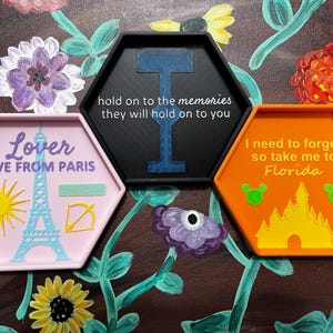 May include: Three hexagonal trays with raised edges. One is pink with a sun, Eiffel Tower, and text that reads "Lover LIVE FROM PARIS". Another is black with a blue design and text: "hold on to the memories they will hold on to you". The last is orange with a castle and text: "I need to forget so take me to Florida".