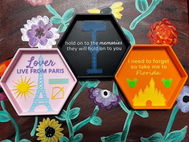 May include: Three hexagonal trays with raised edges. One is pink with a sun, Eiffel Tower, and text that reads "Lover LIVE FROM PARIS". Another is black with a blue design and text: "hold on to the memories they will hold on to you". The last is orange with a castle and text: "I need to forget so take me to Florida".