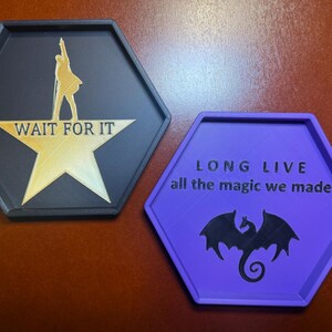 May include: Two hexagonal coasters. One is black with a gold star and the words "WAIT FOR IT" and a silhouette. The other is purple with the words "LONG LIVE all the magic we made" and a dragon silhouette.