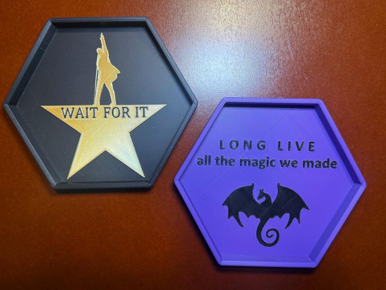 May include: Two hexagonal coasters. One is black with a gold star and the words "WAIT FOR IT" and a silhouette. The other is purple with the words "LONG LIVE all the magic we made" and a dragon silhouette.