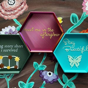 May include: Three hexagonal trays in teal, burgundy, and green, each with a unique inscription. The teal tray says "Stay Beautiful" with a butterfly. The burgundy tray says "meet me in the afterglow". The green tray says "long story short I survived".