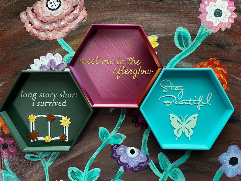May include: Three hexagonal trays in teal, burgundy, and green, each with a unique inscription. The teal tray says "Stay Beautiful" with a butterfly. The burgundy tray says "meet me in the afterglow". The green tray says "long story short I survived".