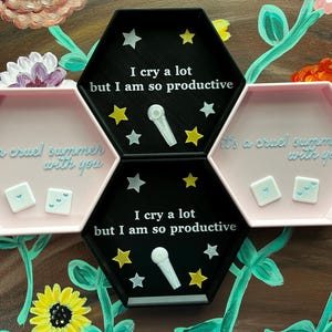 May include: A set of three hexagonal trays. Two are pink and one is black. The black tray has the text "I cry a lot but I am so productive" and star and microphone designs. The pink trays have the text "it's a cruel summer with you" and two small square objects.