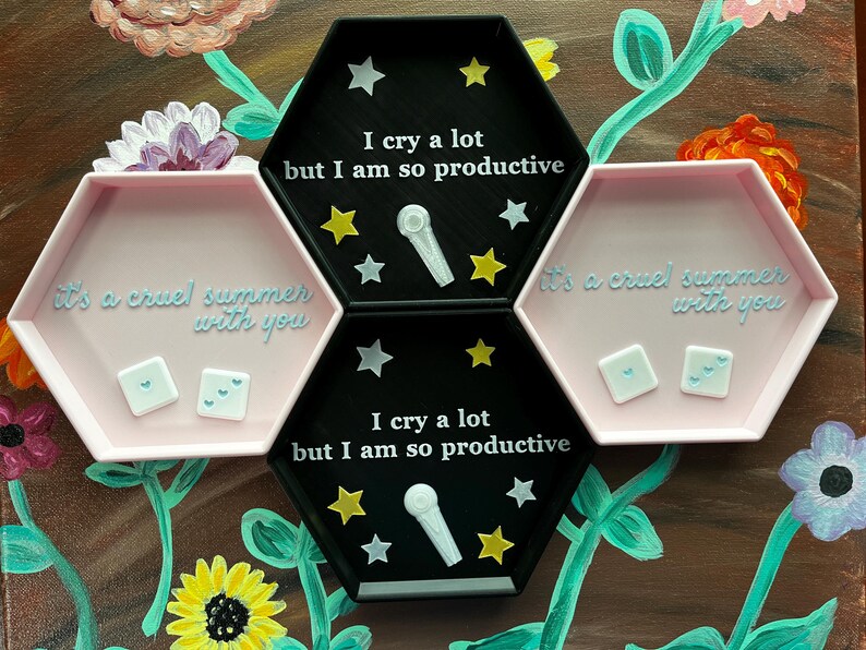 May include: A set of three hexagonal trays. Two are pink and one is black. The black tray has the text "I cry a lot but I am so productive" and star and microphone designs. The pink trays have the text "it's a cruel summer with you" and two small square objects.