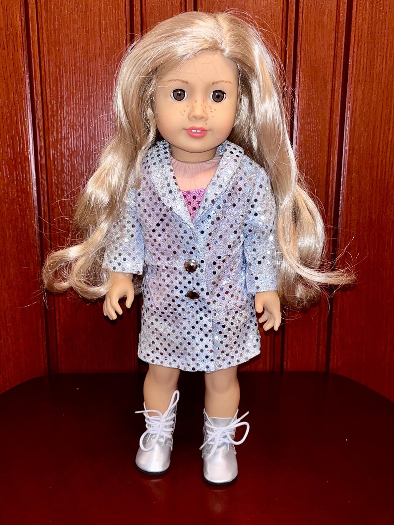 May include: A doll wearing a silver sequined dress and silver boots. The doll has blonde hair and brown eyes.
