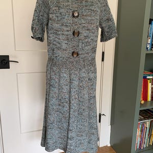May include: A knitted dress in shades of blue, brown, and gray, displayed on a black metal dress form. The dress features short sleeves, a round neckline, and large brown buttons down the front. The skirt flares out from the waist.