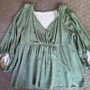 May include: A green floral print top with long sleeves and a V-neckline. The top has a gathered waist and elastic cuffs. The fabric has a small, repeating floral pattern in shades of green and white. The sleeves are long and gathered at the cuffs.