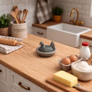 May include: A miniature kitchen scene with a gray hen figurine in a nest, eggs, butter, and a bowl of flour. Wooden utensils and a rolling pin sit near a small potted plant. A white sink and faucet are in the background.