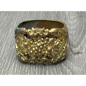 May include: A gold-toned hinged bracelet with intricate floral and grape cluster designs. The bracelet has a detailed, ornate appearance with a chain closure. The metal has a slightly aged patina, adding to its vintage charm.