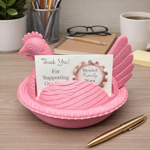 May include: A pink chicken-shaped desk tidy holding a card with the text "Thank You! For Supporting Our..." The organiser has a detailed design and a gold pen rests in front of it. The card also has the text "Blended Family Worx".