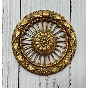 Puede incluir: Un tirador de armario circular dorado, con un centro floral y un diseño de radios. El tirador está rodeado por un borde detallado de hojas y bayas. Es probable que el herraje sea de metal.