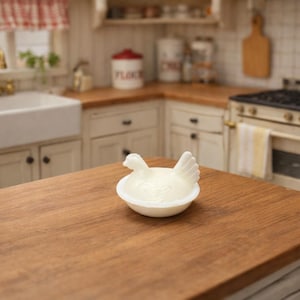 May include: A white ceramic chicken-shaped butter dish sits on a wooden countertop in a kitchen setting. The kitchen features white cabinets, a farmhouse sink, and a vintage stove. Red and white checkered curtains are visible in the background.