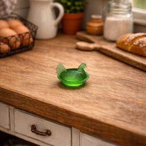 May include: A small, green glass hen-shaped dish sits on a wooden countertop in a kitchen setting. The dish is detailed with a textured design. Other kitchen items include a wire basket of eggs, a white pitcher, and a loaf of bread.