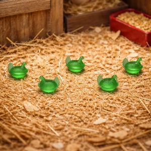 May include: Five small, translucent green chicken figurines are scattered on a bed of straw. The figurines have a detailed wing design. In the background, a wooden structure with eggs and feed is visible, suggesting a farm or poultry theme.