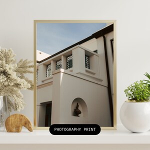 May include: A photograph of a white building with a balcony and a decorative archway. The building has a modern design with clean lines and a minimalist aesthetic. The photograph is taken from a low angle, giving the viewer a sense of perspective. The building is set against a clear blue sky.