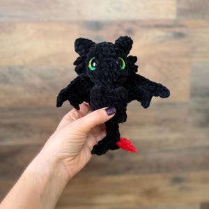 Può includere: Un peluche a forma di drago nero all'uncinetto con occhi verdi e coda rossa. Il drago ha le ali ed è tenuto in mano. Il peluche è realizzato con un filato morbido e strutturato.