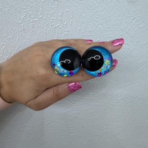 May include: Two round, blue, domed cabochons featuring large, solid black pupils and embedded holographic star-shaped glitter. These circular craft eyes have a vibrant, reflective finish with a clear, smooth surface texture and a bright blue iris.
