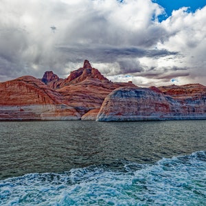 May include: A view of red rock formations rising from a body of water. The water is calm and blue, with white foam from a boat's wake in the foreground.