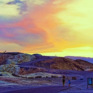 May include: A couple walks along a path in Death Valley National Park, with colorful rock formations and a vibrant sunset sky in the background.