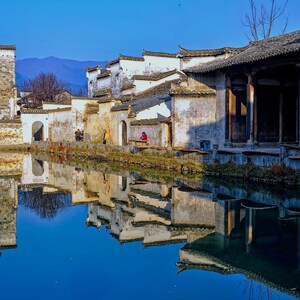 Peut inclure: Vue d'un village traditionnel chinois avec des bâtiments blanchis à la chaux et un bassin d'eau réfléchissant. Les bâtiments ont des toits en tuiles et sont entourés d'un mur de pierre. L'eau reflète les bâtiments et le ciel.