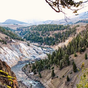 May include: A scenic landscape featuring a river winding through a canyon. The canyon walls display shades of brown and white, with patches of green trees. The sky is overcast, and the overall scene evokes a sense of natural beauty and tranquility.