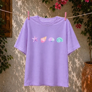 Puede incluir: Una camiseta de manga corta de color morado claro con una fila horizontal de cuatro ilustraciones de conchas marinas en el pecho. El gráfico incluye una estrella de mar rosa, una concha moteada, una vieira en forma de abanico y un caracol marino.