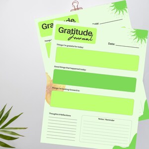 Puede incluir: Una pila de páginas de diario de gratitud verde claro con un encabezado verde lima que dice "Gratitude Journal". Las páginas tienen secciones para reflexiones diarias, incluyendo indicaciones de gratitud, cosas buenas y metas futuras. Un clip de papel dorado sujeta las páginas.