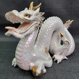May include: A white porcelain dragon figurine with gold accents. The dragon is curled up with its head raised and its mouth open. The figurine has a pearlescent finish.