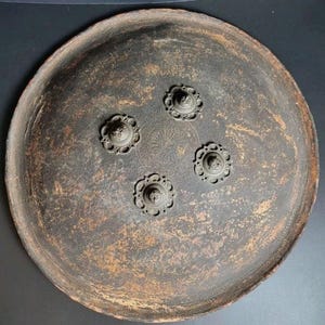 May include: A circular, dark-toned metal plate or tray with a weathered, textured surface. The center features four symmetrical, raised floral-shaped medallions arranged in a square pattern, surrounding a small, finely engraved circular sunburst motif.