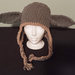 Pode incluir: Um chapéu de crochê marrom e bege com abas de orelha longas e um tassel trançado. O chapéu está em uma cabeça de manequim.